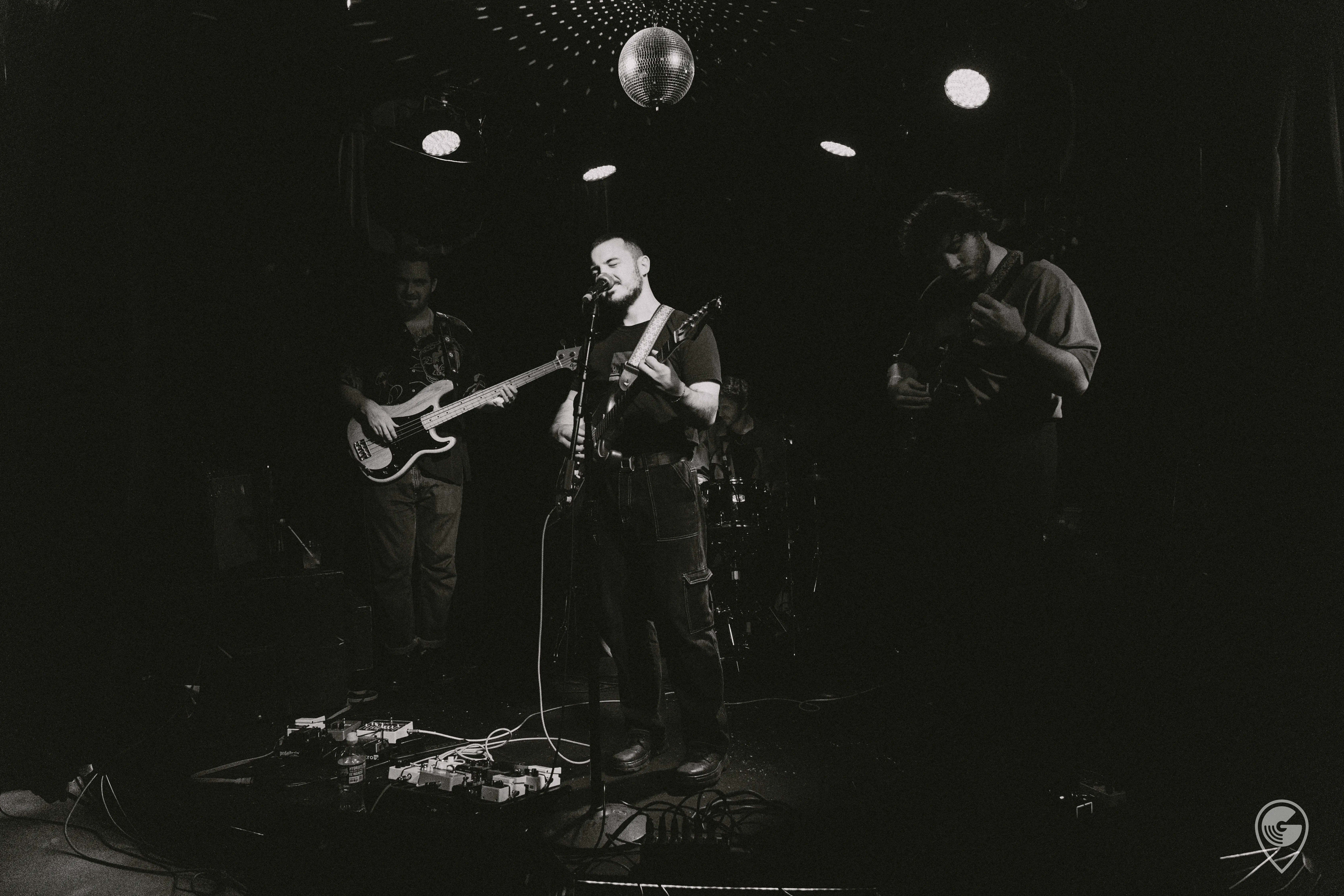 A three-piece band performs on a dimly lit club stage in black and white — a singer plays guitar at the center mic, flanked by a bassist and a guitarist, beneath a disco ball and stage lights