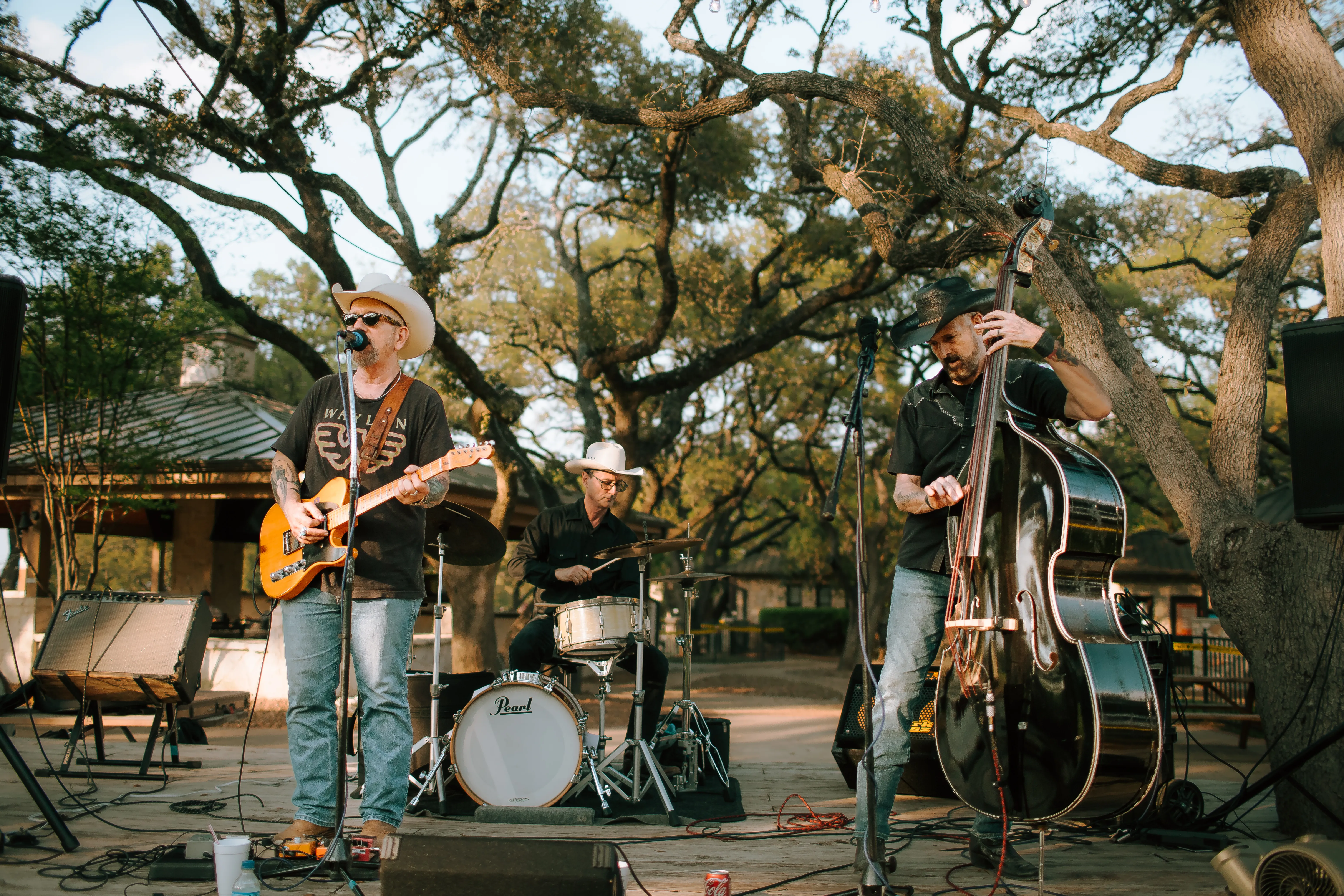 Country trio plays under a live oak at golden hour: a guitarist-singer and upright bassist in cowboy hats, with a drummer
