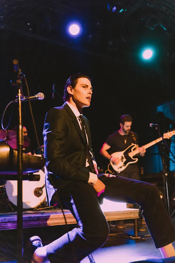 A vocalist in a suit performing on a dimly lit club stage, with a bassist and drummer playing behind him