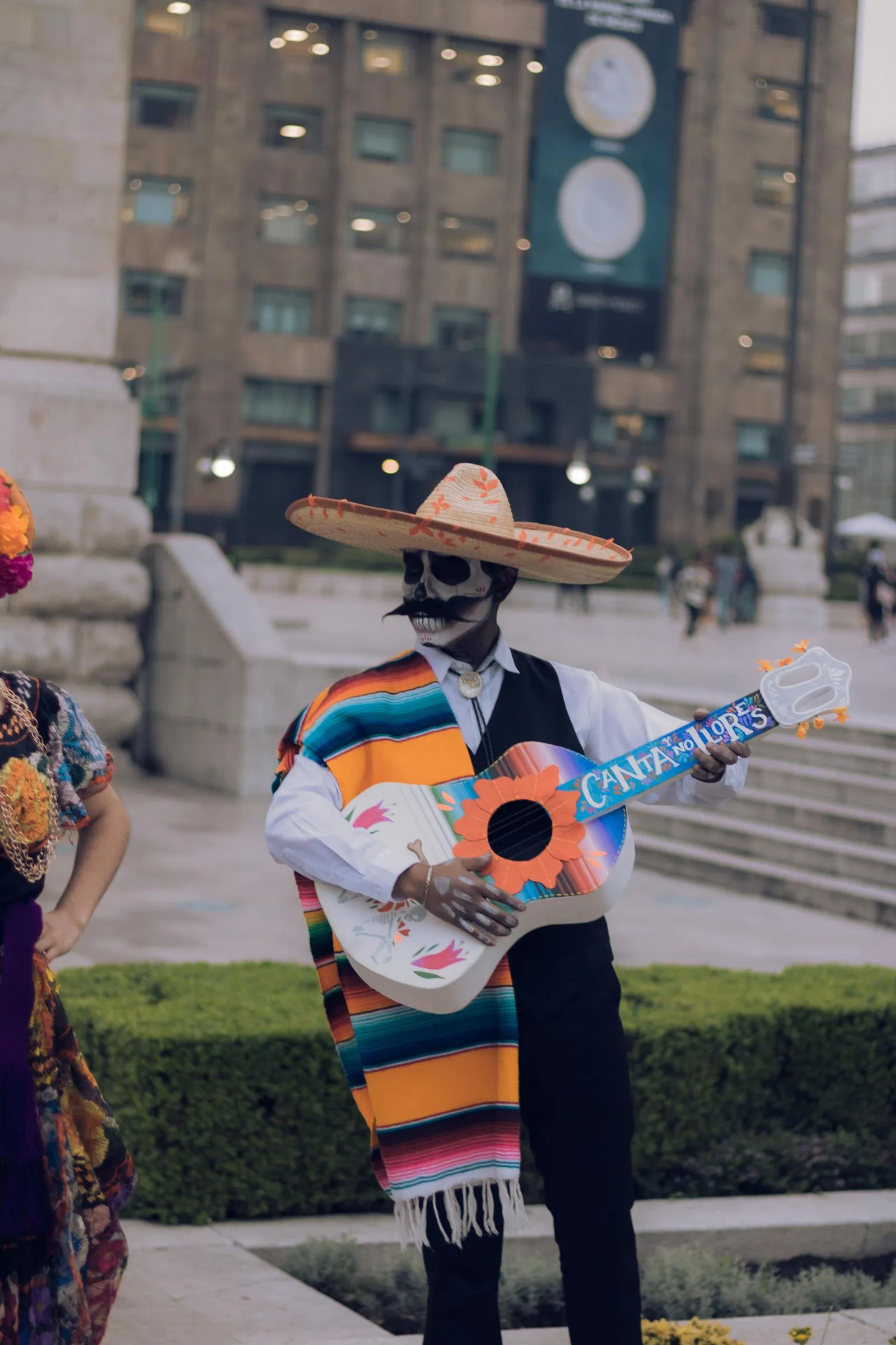 A street performer in Día de los Muertos calavera face paint, a wide-brimmed sombrero, and a striped serape plays a hand-painted guitar during a public celebration