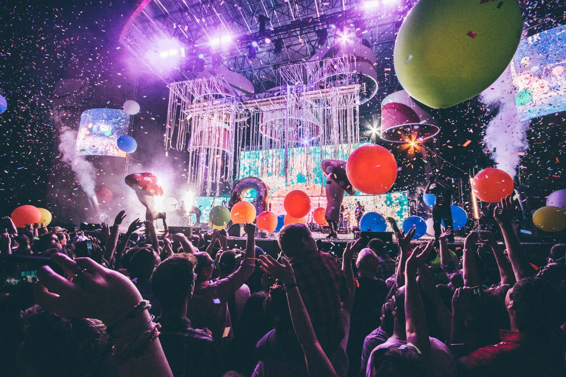 A packed crowd celebrates in front of a brightly lit stage, hands in the air amid falling confetti and balloons at a live music event