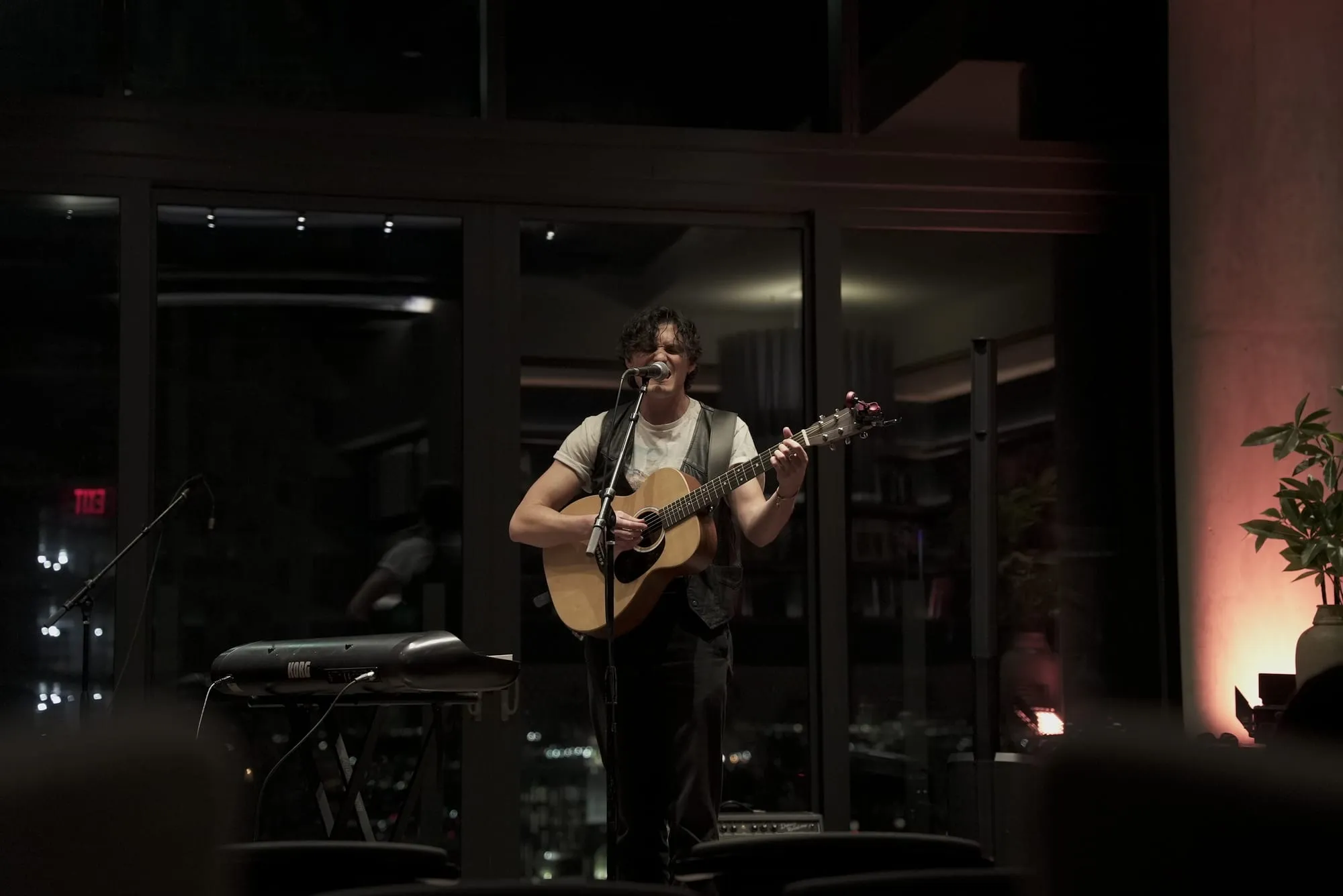 A solo musician sings and plays acoustic guitar during a live set in a dimly lit hotel lounge, framed by floor-to-ceiling windows and warm red light