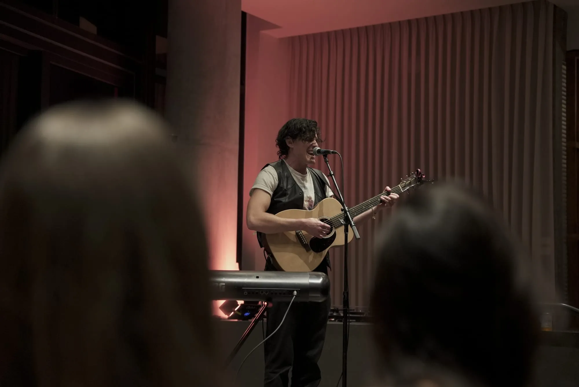A live acoustic performance seen from the back of a hotel lounge, over the heads of a seated audience, the musician lit against a warm red backdrop as the room watches