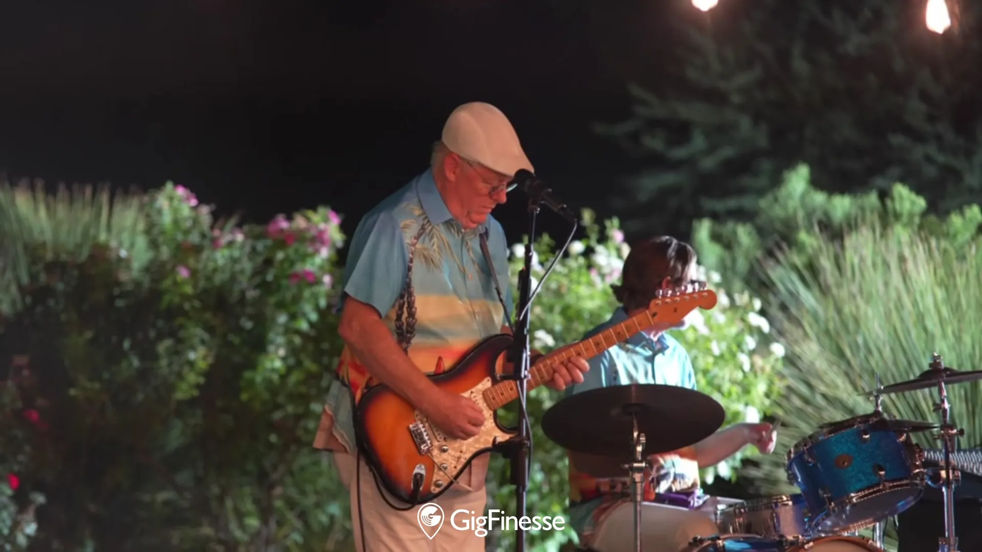 A live band performs outdoors at an Invited Clubs member event at night — a singer-guitarist at the mic, with a drummer behind him, framed by garden lighting