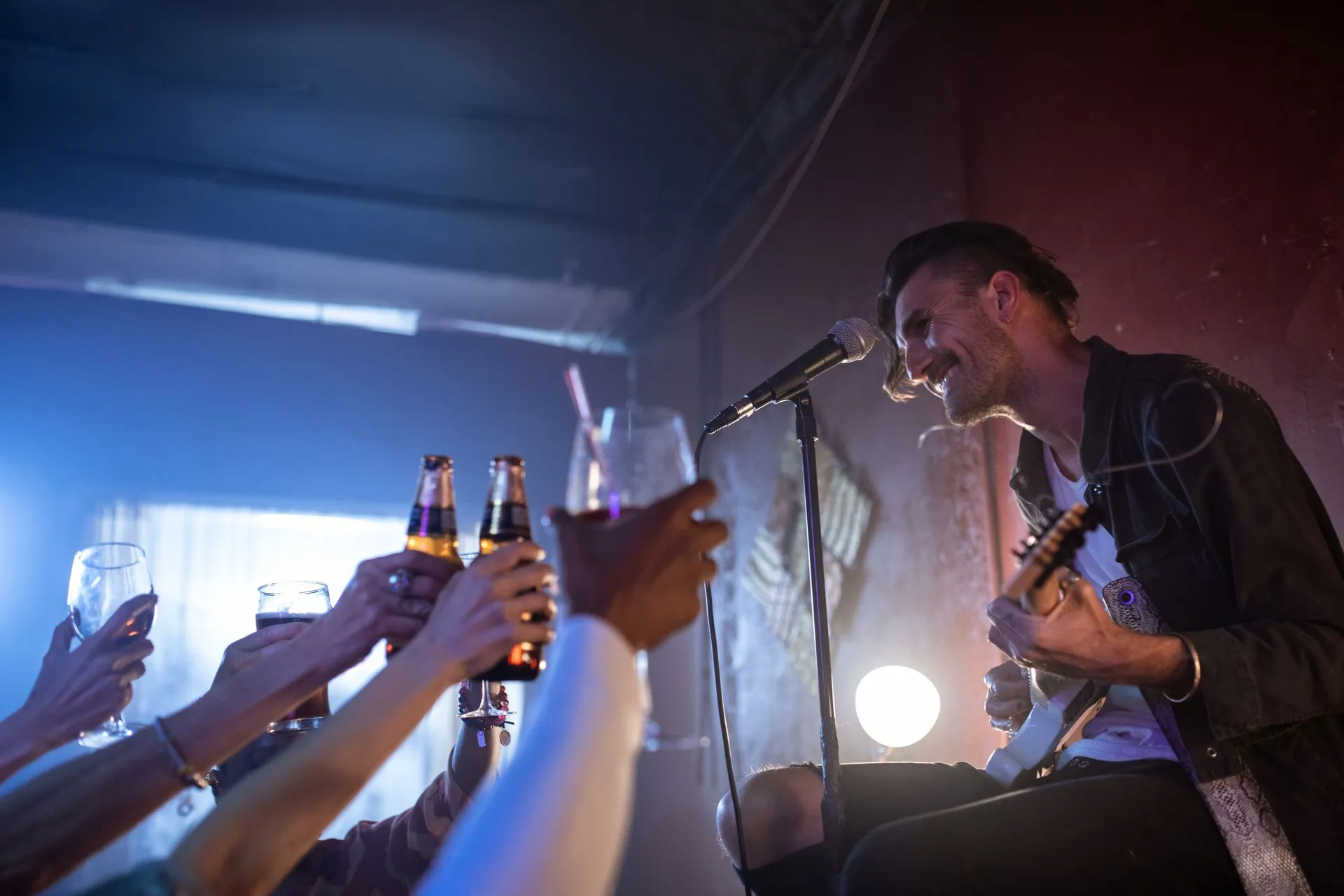 A solo musician sings into a mic and plays guitar in a dimly lit venue while guests in the foreground raise beer bottles and glasses in a toast