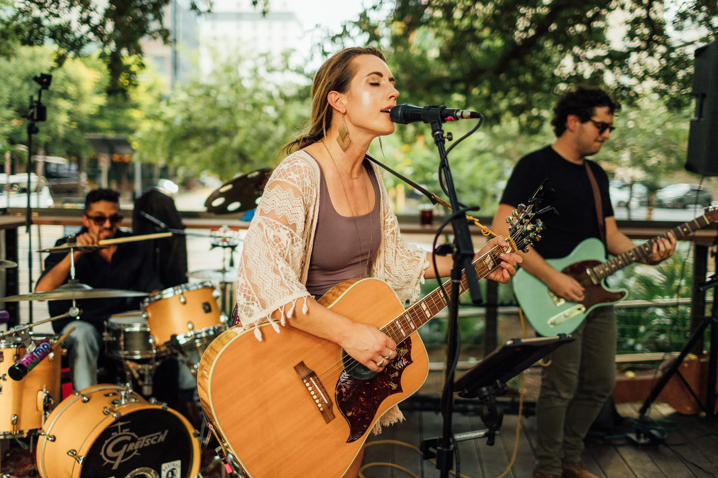 A trio performs live on an outdoor patio at a bar: a woman sings and plays acoustic guitar at the mic, with a drummer and an electric guitarist beside her