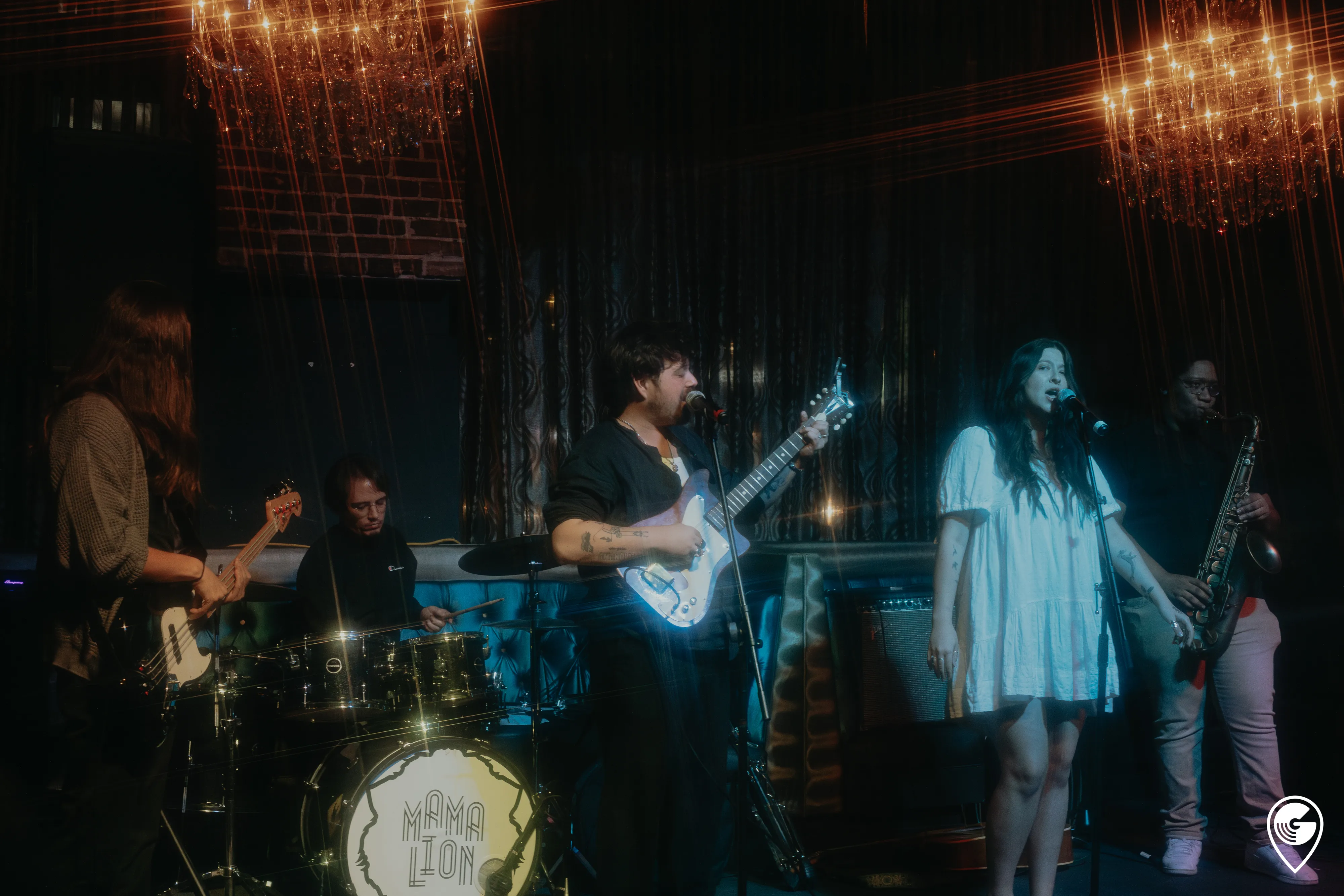 A five-piece band performs on an intimate, chandelier-lit lounge stage with two vocalists, sax, guitar, bass, and drums