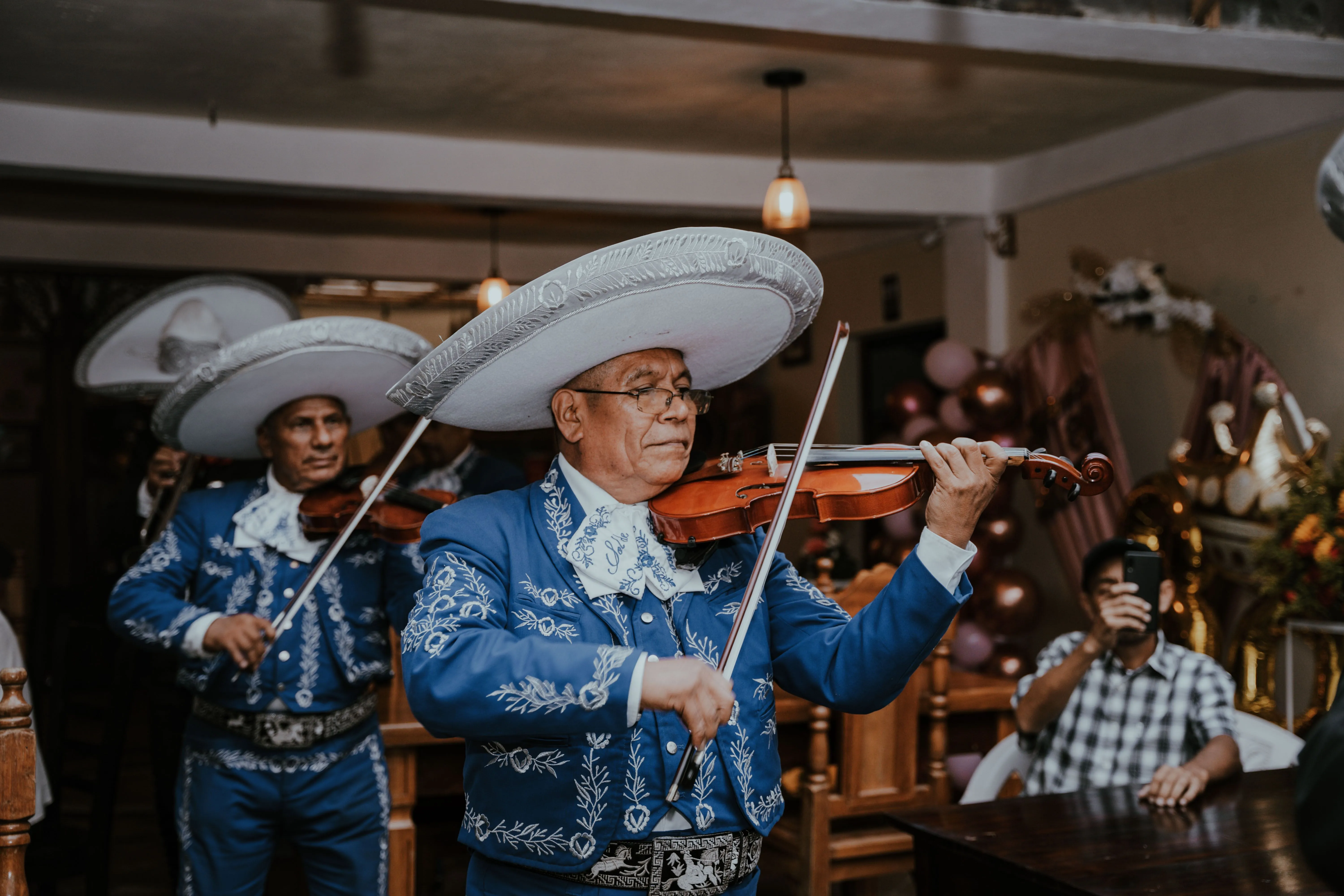 Mariachi musicians in blue embroidered charro suits and white sombreros play violins during a live performance indoors