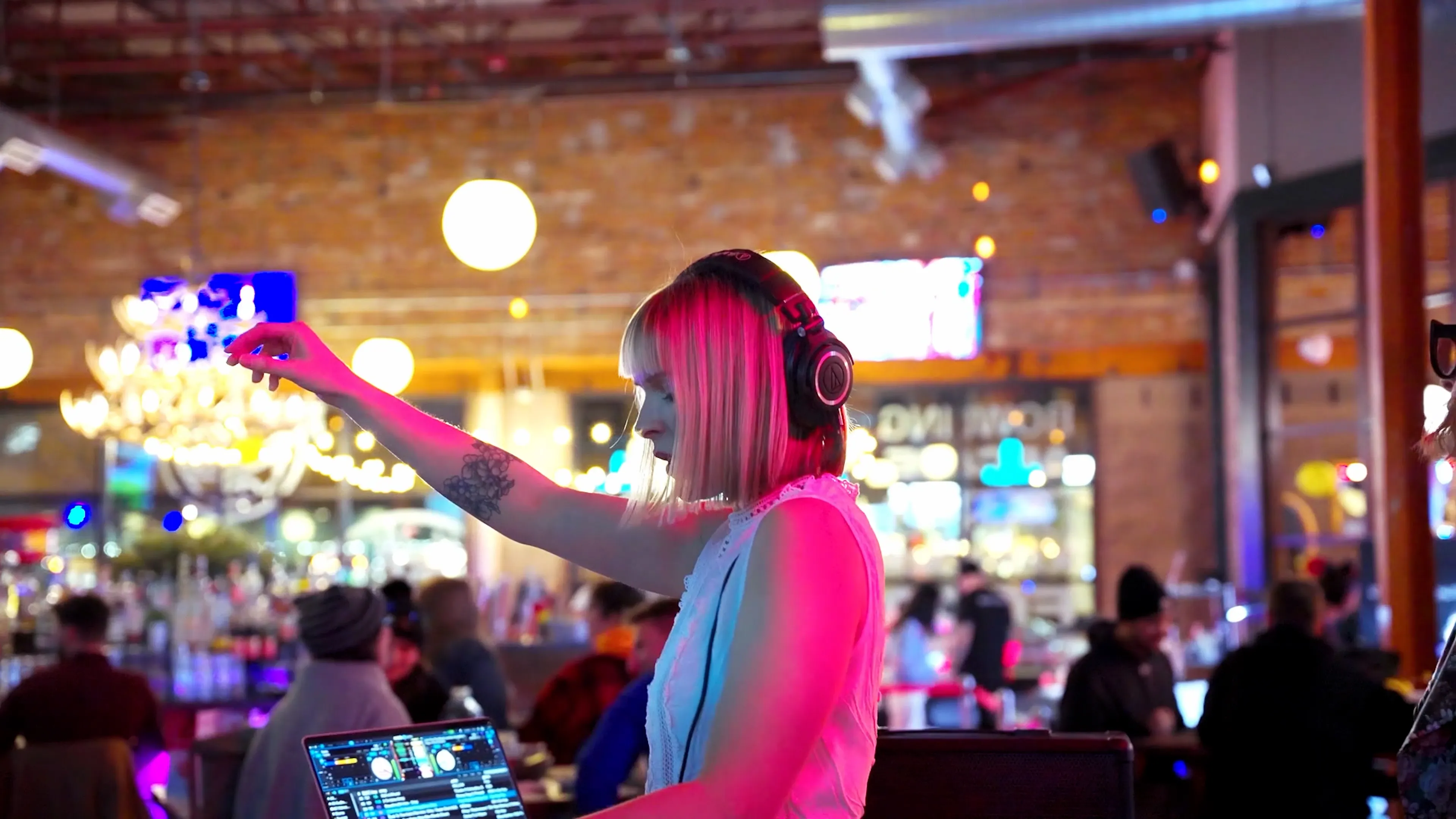 A DJ spins a live set at Punch Bowl Social in Denver, backlit by warm chandelier light above a busy bar crowd