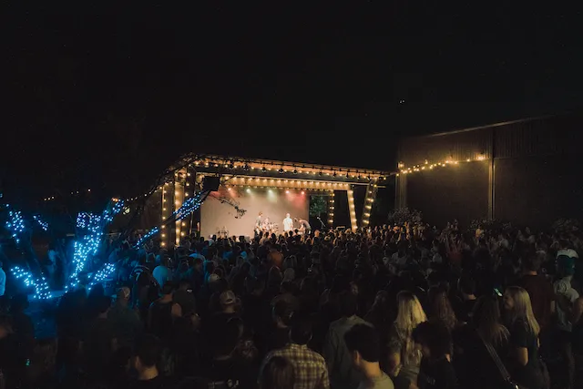 A crowd gathered before a brightly lit outdoor stage at night, framed by string lights, at a live music event